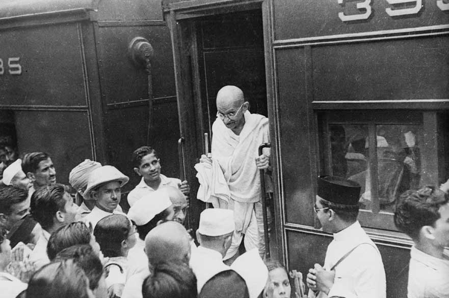 A crowd gathers to welcome Mahatma Gandhi as he arrives by train at a railway station.
